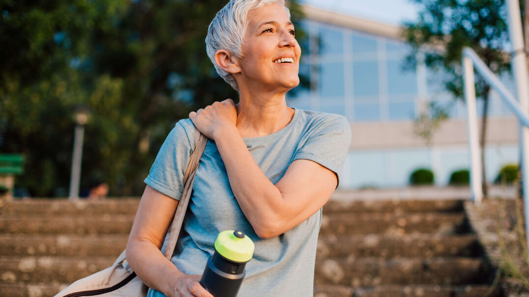 A woman smiles in sunshine. She is carrying a water bottle, suggesting she has been exercising.